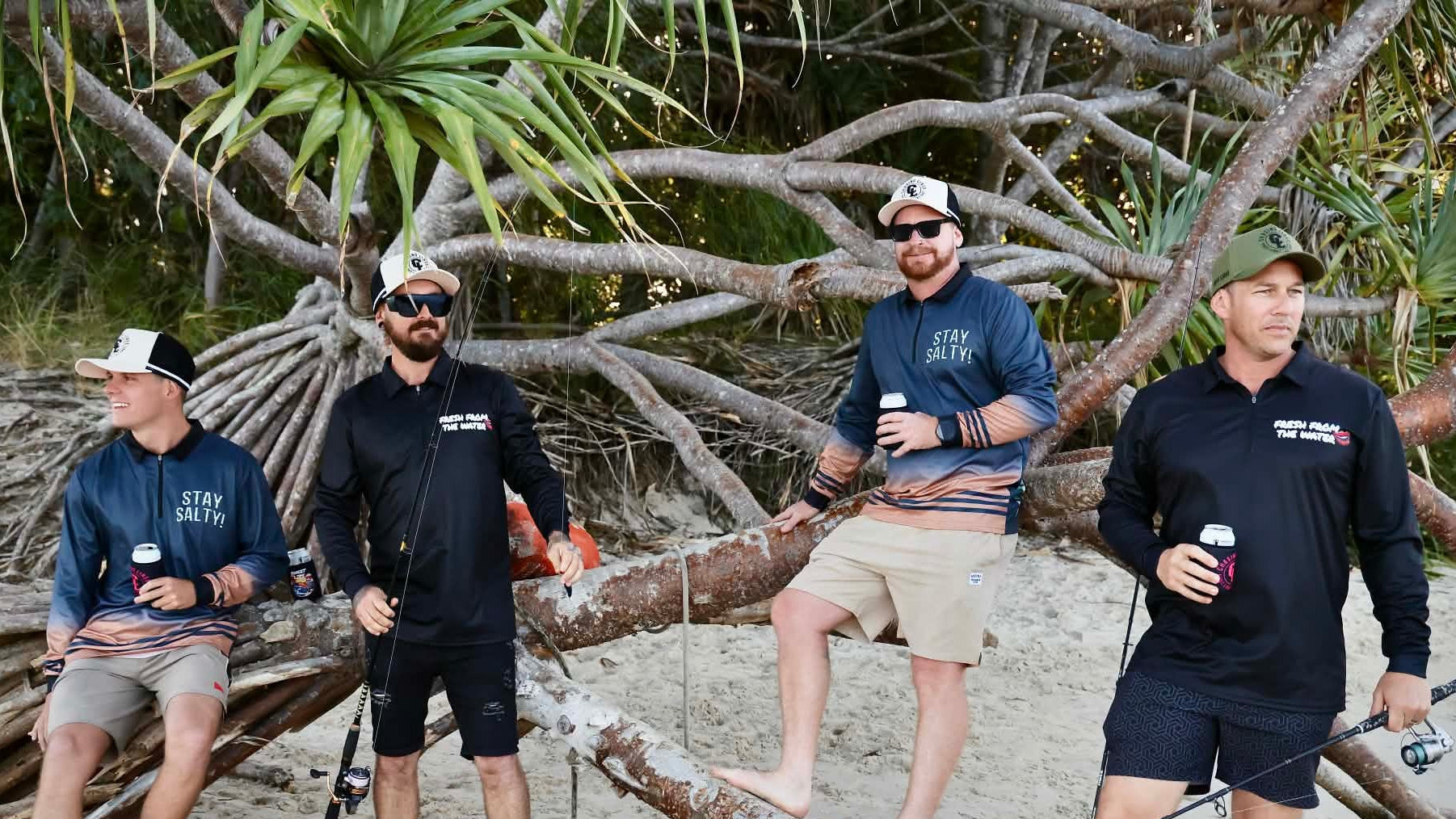 Four men on a beach with fishing equipment, surrounded by large tree roots and greenery.